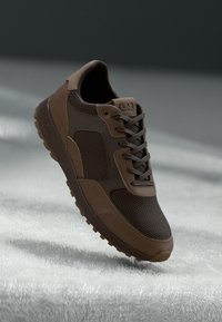 Brown athletic shoe featuring a mesh upper and leather accents, with a textured sole and round laces. The design is low-top with branding.