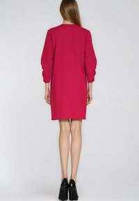 Kobieta ubrana w fuksjową sukienkę do kolan z rękawami do łokcia, stojąca tyłem, w czarnych szpilkach.