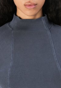 Pogled od blizu na osebo, ki nosi rebrast, visok ovratnik, dolg rokav sivi zgornji del, ki prikazuje izrez in ustnice.