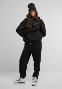 Haut à capuche noir surdimensionné avec poche kangourou, joggers assortis, bonnet noir en tricot texturé et baskets blanches avec accents verts.