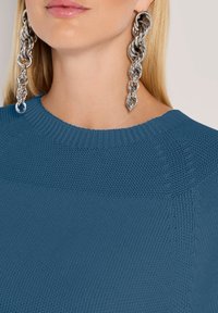 Nahaufnahme einer Person mit übergroßen silbernen Kettenohrringen und einem blauen strukturierten Strickpullover mit geripptem Ausschnitt.