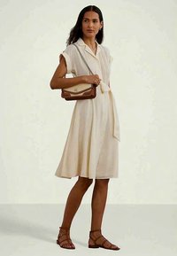 Ženska v kremastih oblekah s frontnimi gumbi in pasom, drži rjav nahrbtnik in nosi rjav sandale s trakovi.