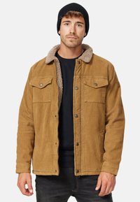 Katoenen jack in tan met een faux shearling kraag, drukknoopsluitingen, twee borstzakken en ribbelmanchetten. Zwarte beanie op het hoofd.