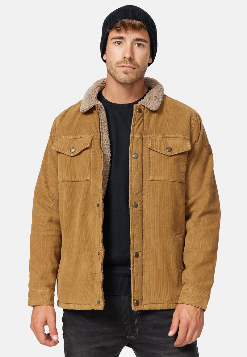 Katoenen jack in tan met een faux shearling kraag, drukknoopsluitingen, twee borstzakken en ribbelmanchetten. Zwarte beanie op het hoofd.