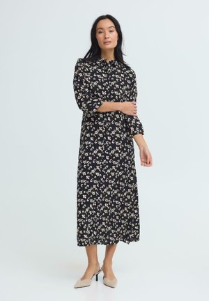 Kobieta stojąca, ubrana w czarną, długą sukienkę w kwiaty z rękawami 3/4 oraz beżowe, szpiczaste szpilki, na tle jednolitego jasnego tła.