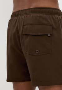 Braune gewebte Shorts mit elastischem Bund, ausgestattet mit einer Gesäßtasche mit Klappe und einem Ösen-Detail. Strukturierte Stoffoberfläche.