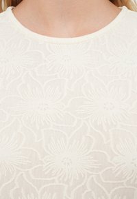 Λευκή μπλούζα με φλοράλ σχέδιο σε υφασμάτινη υφή, με στρογγυλή λαιμόκοψη και διακριτικές ανάγλυφες λεπτομέρειες λουλουδιών σε όλη της την επιφάνεια.