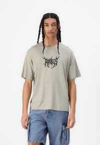 T-shirt verde oliva a maniche corte, con un logo grafico nero. Abbinato a pantaloni cargo blu jeans, che mostrano una vestibilità rilassata.