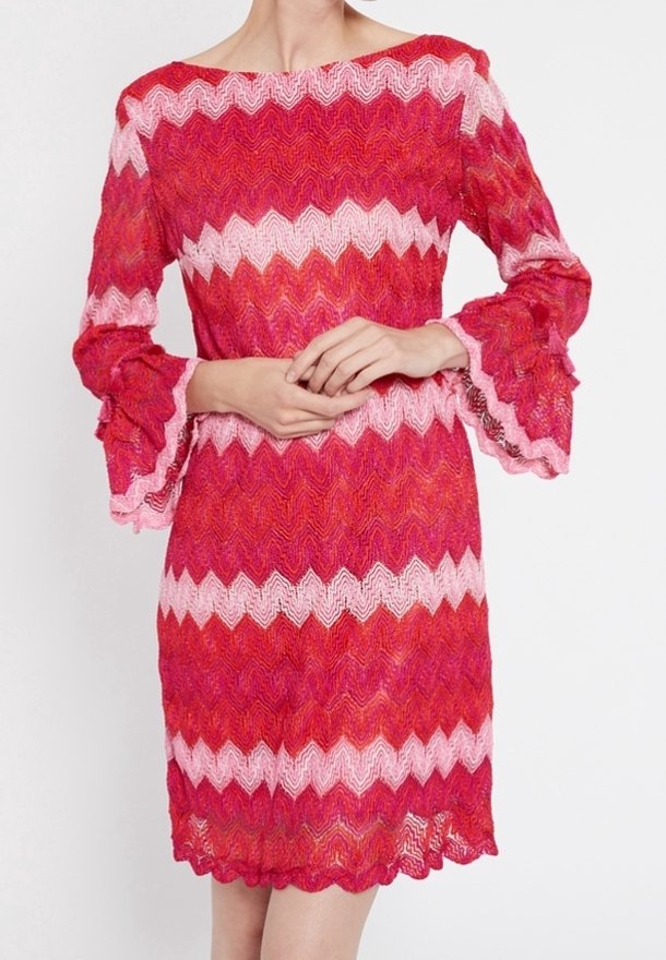 Femme portant une robe tricotée rose et rouge à motif zigzag, avec de longues manches évasées et un ourlet festonné, debout, les mains jointes.