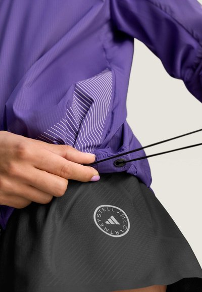 Chaqueta deportiva morada con un diseño a rayas, que cuenta con un ajuste de cordón. Parte inferior de falda negra con un logo en un patrón circular.