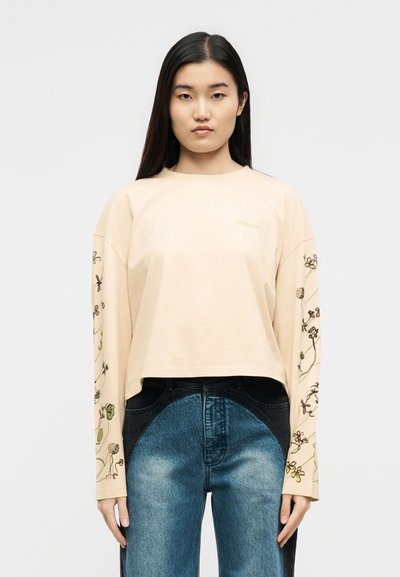 Γυναίκα με μπεζ crop top μακρυμάνικο με κεντήματα με λουλούδια στα μανίκια και μπλε δίχρωμα τζιν, στέκεται μπροστά σε απλό φόντο.