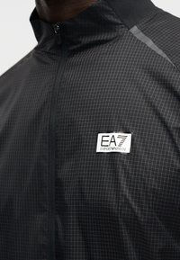 Μαύρο ελαφρύ μπουφάν με πλέγμα σχεδίασης, ψηλό γιακά και φερμουάρ. Διαθέτει ετικέτα με λογότυπο EA7 Emporio Armani στο στήθος.