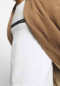 Brązowy sweter o teksturowanym wykończeniu, nałożony na białą koszulę z poziomymi paskami w jasnym żółtym i granatowym kolorze.