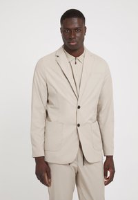 Blazer bej ușor, cu un design ajustat, două buzunare frontale, nasturi negri și o textură netedă, purtat peste o cămașă bej asortată.