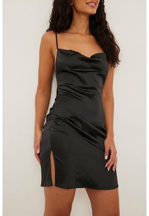 Kobieta w czarnej satynowej mini sukience na cienkich ramiączkach z bocznym rozcięciem, stojąca na tle jasnego, jednolitego tła.