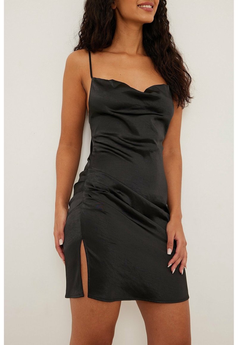 Femme portant une mini robe en satin noire à fines bretelles et fente latérale, debout devant un fond clair uni.