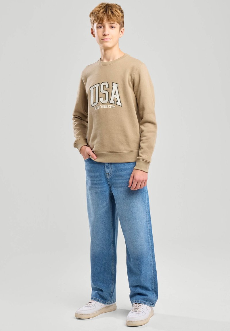 Beige sweatshirt with "USA NEW YORK CITY" printed in gray, paired with light blue baggy jeans and white sneakers. Smooth fabric, casual design.