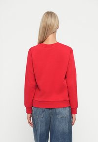 Felpa rossa con colletto tondo e polsini a costine, realizzata in materiale morbido. Abbinata a jeans in denim blu, vista di spalle.