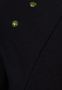 Tessuto lavorato a maglia nero con un motivo testurizzato, caratterizzato da tre bottoni decorativi verdi lucidi posizionati strategicamente sulla superficie.