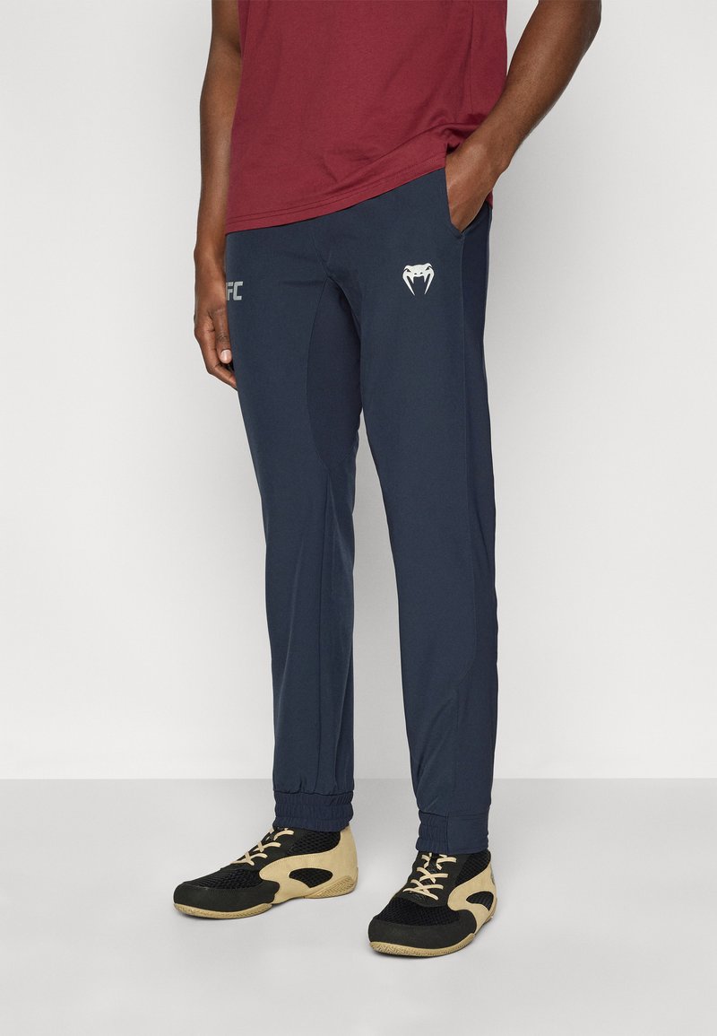 Παντελόνι αθλητικό σε ναυτικό μπλε με εφαρμοστή γραμμή, ελαστικές μανσέτες και λογότυπο UFC· φοριέται με αθλητικά παπούτσια σε μαύρο και εκρού χρώμα και μπλούζα μπόρντο.