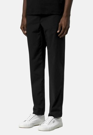 Uomo che indossa pantaloni neri a gamba dritta e sneakers bianche con lacci, in piedi davanti a uno sfondo chiaro e uniforme.