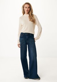Ljusbeige långärmad tröja med muddkrage, kombinerad med mörkblå vida jeans. Jeansen har en klassisk femficksdesign.