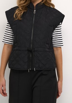 Frau trägt eine ärmellose, schwarz gesteppte Weste mit Frontreißverschluss und Gürtel über einem schwarz-weiß gestreiften Kurzarmshirt und schwarzen Hosen.