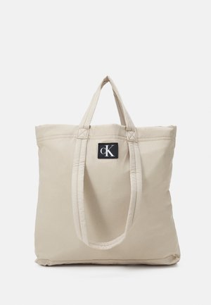 Borsa a tote in tessuto beige con due manici e una toppa quadrata nera con il logo bianco "cK" sul davanti.
