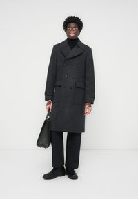 Manteau en laine gris foncé avec un design croisé, larges revers et deux poches avant. Associé à un col roulé noir et un pantalon.