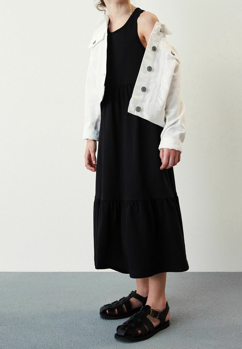 Personne portant une robe midi noire sans manches, une veste blanche boutonnée tombant des épaules, et des sandales noires à lanières, debout sur un sol gris.
