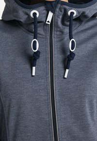 Veste grise à fermeture éclair avec un col montant, dotée d'une capuche à cordon de serrage et d'accents de quincaillerie blancs. Confectionnée en tissu lisse et texturé.