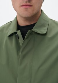 Chaqueta ligera verde con cuello, que presenta un cierre delantero de botones y una textura suave. Lleva puesta sobre una camisa negra.