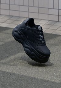 Fekete sportcipők vastag talppal, sima bőr felső résszel és texturált gumitalppal. Felszereltségük zsinórrendszert és logós részleteket tartalmaz.