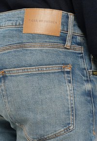 Light wash denim jeans with orange stitching, leather tag reading "TIGER OF SWEDEN," and standard back pocket design.