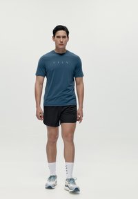 Košulja kratkih rukava plave boje s sivozelenim akcentima, u kombinaciji s crnim šortsevima. Ima laganu tkaninu i sportski dizajn, idealna za treninge.