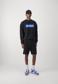 Μαύρη φούτερ με το λογότυπο "HUGO" σε έντονο μπλε χρώμα, συνδυασμένη με μαύρες βερμούδες. Μαύρο καπέλο, λευκές κάλτσες και μπλε/λευκά αθλητικά παπούτσια. Απλός σχεδιασμός.