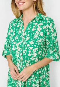 Grön blommig klänning med vita mönster, korta ärmar, krage, knappframsida och samlad midjedetalj. Mjuk, texturerad tyg.