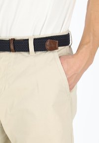 Beige chinot, jossa on kudottu tummansininen vyö, jossa on ruskea nahkaaccentti; sileä kangas ja sivutaskut näkyvissä.