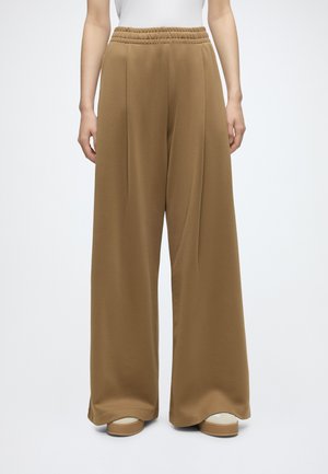 Femme portant un pantalon marron ample à jambes larges avec une ceinture élastique, debout droite, pieds légèrement écartés, chaussures blanches partiellement visibles.