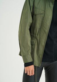 Olive green jacket with a relaxed fit, featuring a chest pocket, elastic cuffs, and a visible brand logo on the sleeve. Smooth texture.