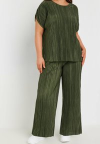 Grön plisserad topp och vida byxor, tillverkad av ett glänsande material. Outfitten har en lös passform och korta ärmar, i kombination med vita skor.