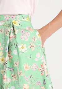 Jupe fleurie en tissu vert menthe avec une finition texturée. Elle présente une taille haute, une poche latérale et des motifs floraux multicolores de différentes tailles.