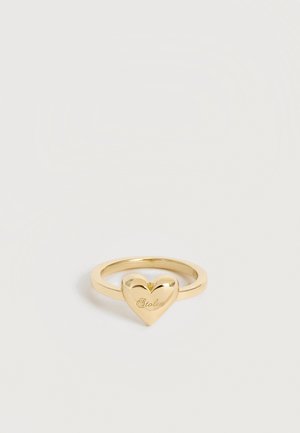 Anillo de oro con un charm pulido en forma de corazón grabado con la palabra "Stolen" sobre un fondo blanco liso.