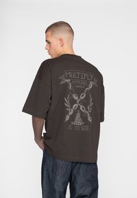 Mørkebrun oversized T-shirt med et grafisk design på bagsiden, der viser "MULTIPLY APPAREL," et symbol og teksten "ALL YOU NEED."