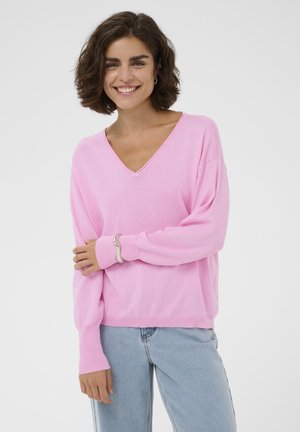 Femme souriante aux cheveux bruns courts, portant un pull rose à col en V et un jean bleu clair, debout devant un fond uni.