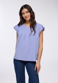 Blusa viola chiaro con maniche corte a volant, scollatura rotonda e un delicato dettaglio plissettato al centro. Abbinata a pantaloni in denim scuro.