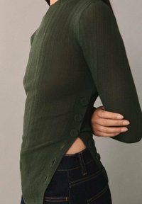 Zielona, prążkowana bluzka z długim rękawem i wysokim kołnierzem, ozdobiona guzikami po lewej stronie, zestawiona z ciemnoniebieskimi dżinsami.