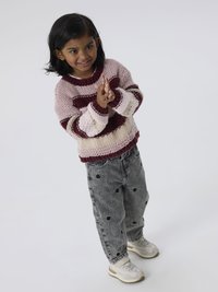 Camisola de malha multicolorida com riscas rosa, bordô e creme. Jeans cinzentos com buracos pretos. Ténis brancos com detalhes em cinza.