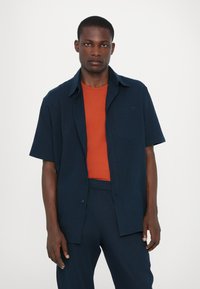 Kurzarm-Button-Down-Hemd in Marineblau über einem roten Rundhals-T-Shirt. Das Hemd hat eine aufgesetzte Tasche und ein dezentes Logodesign.