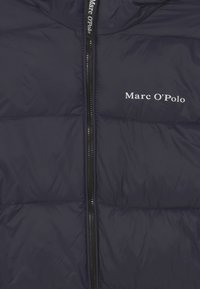 Marinblå puffjacka med kviltad textur, hel dragkedja och vit logotyp "Marc O'Polo" på vänster bröst. Slätt, lättviktigt tyg.